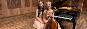 Two young women, one holding a cello, sitting on a piano stool together in front of a grand piano