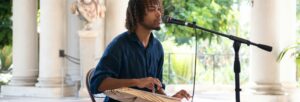 A young man in a blue shirt, seated before a microphone and holding an inanga (East African zither)