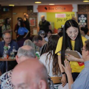 A young woman with long dark hair and a yellow tshirt talking to a man in a blue shirt in a crowded foyer. You can see the Box Office sign in orange behind her