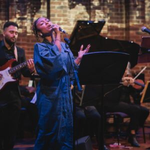 A woman in a blue robe singing into a microphone on stage with music stands. A man with an electric guitar plays behind her.