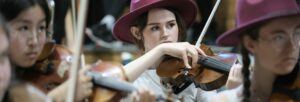A group of young people wearing burgundy trilby hats playing stringed instruments