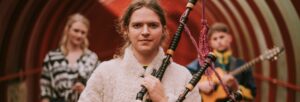 A person with brown hair tied back, holding bagpipes. Out of focus behind them are two more musicians