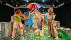 four women on a stage, two playing violins, one opening a rainbow striped umbrella and scattering coloured confetti on the fourth woman who is laughing