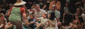 An audience of excited children and adults smiling at a performer on stage wearing a hat
