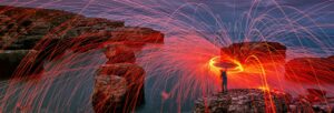 A silhouetted figure stood on a cliff over water wielding bright orange and red sparks in a circle over their head