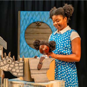 A young black woman in a blue polka dot dress smiling down at a little puppet of herself