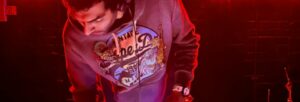 A dark haired man in a hooded top, dimly lit by red light, bends forward over a set of decks
