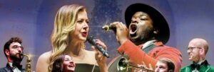Two vocalists, one female and one male, sing in front of a Christmas tree surrounded by musicians playing different instruments.