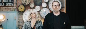 A woman with long blonde hair standing next to a man with dark hair, beard and glasses in front of a wall of clocks