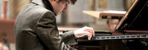 A man with dark curly hair in a smart jacket playing a grand piano