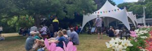 People gathered, some seated, some milling about, outdoors in a summer garden with a marquee and bunting