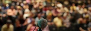 A microphone points to the camera with a blurring view of an audience beyond.