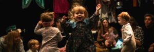 Small children playing, with particular focus on a little girl with red hair smiling with her hands held up in the air