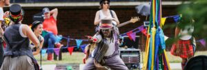 Man with a top hat, colourful waistcoat, checked trousers and colourful socks reaching out to an audience of families with musicians and a maypole in the background