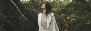 A dark haired woman in a white jacket and top stood in woodland