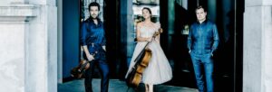 A man wearing dark blue holding a violin stood next to a woman wearing a pale grey tulle dress with a cello. To her other side, a man wearing dark blue stands with his hand in his pockets.