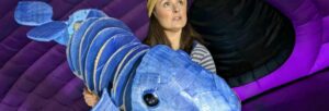 A woman wearing a yellow beanie holding a large puppet of a whale made of cardboard and paper mache