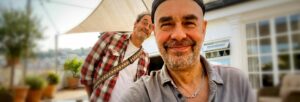 A smiling man in a black hat close to the camera with a man in a check shirt behind him smiling over his shoulder