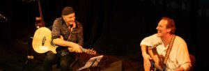 Two white men playing guitars, sat on stage whilst laughing
