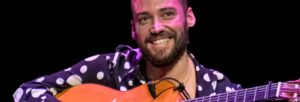 Man holding guitar on stage smiling under stage lighting.