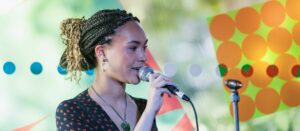 A young Black women in a dark shirt singing into a microphone, with a window showing trees and colours behind her.
