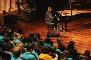 A man seated at a Steinway grand piano smiles at his audience of school children whilst he plays
