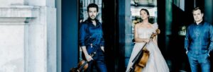 A man wearing dark blue holding a violin stood next to a woman wearing a pale grey tulle dress with a cello. To her other side, a man wearing dark blue stands with his hand in his pockets.