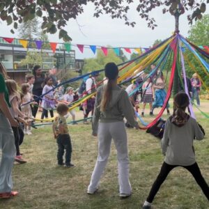 People stood round a Maypole holding string under colourful bunting and trees.