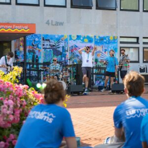 A young band perform outdoors before a brightly coloured artwork. Two young people in blue Turner Sims tshirts watch