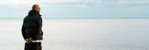 A man wearing a dark puffy jacket and dark trousers staring out across a calm sea to the horizon