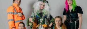 A man in high viz clothing, a man with a worried expression, a woman with green and white curls, elaborately dressed in green and leaves, a woman with long red hair and a woman in a black dress with half pink and half green hair all staring at the camera
