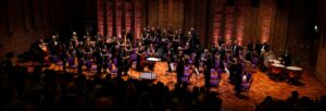 An orchestra on their feet with pink and orange lights around the stage.