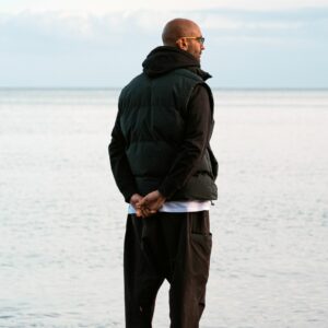 A man wearing a dark puffy jacket and dark trousers staring out across a calm sea to the horizon
