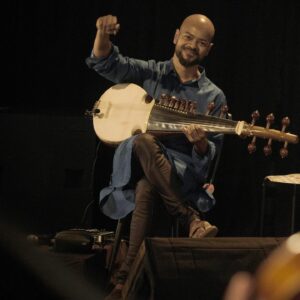 An Indian man in a blue tunic seated and playing the sarod