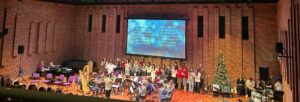 Image of a stage with musicians on, with Christmas lights and a Christmas tree to the right.