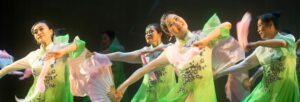 Multiple women dressed in green floaty costumes saving on a stage.