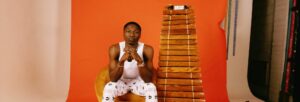 One black man looking at the camera in front of an orange background. With a large wooden instrument to his side.