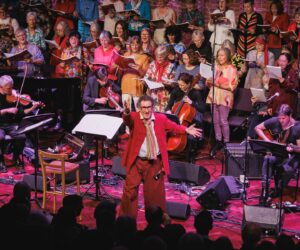 A man in a red suit in front of a large mixed choir facing an applauding audience