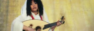 A woman holding a string instrument with white fabric draped over her head and shoulders.