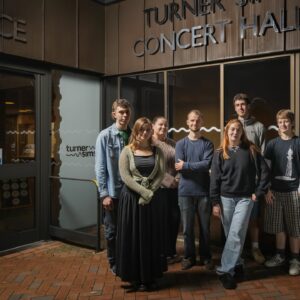 7 young people stood outside at night-time in front of a sing that reads 'Turner Sims Concert Hall'